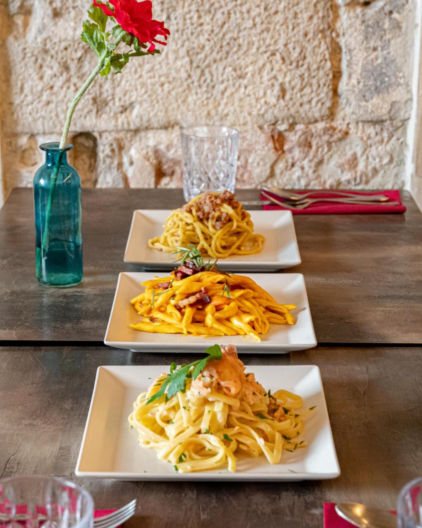 Tre piatti di pasta in fila sul tavolo di legno davanti al muro in pietra, con una rosa nel vaso