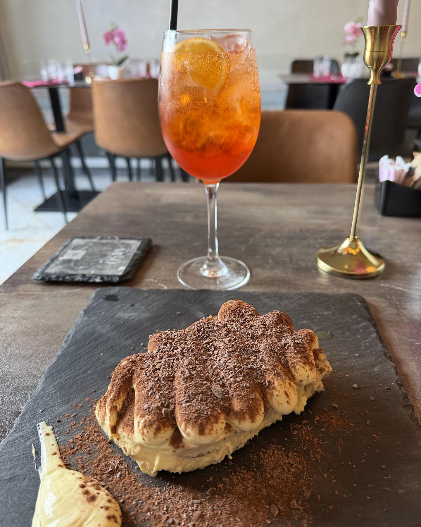 Tiramisù classico spolverato di cacao accanto a un Aperol spritz, sul tavolo di legno