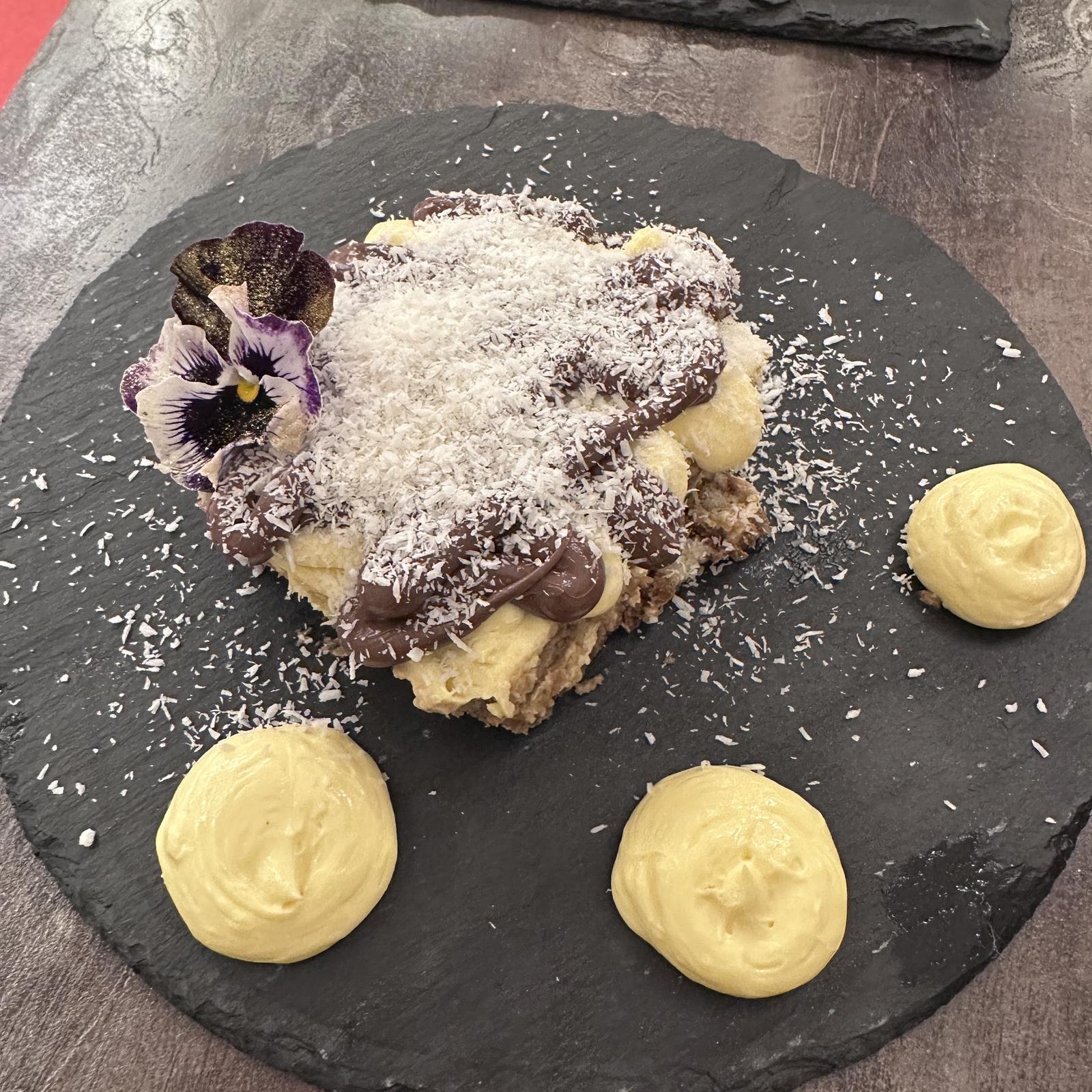 Tiramisù al cocco con una viola del pensiero e tre ciuffi di crema sul piatto d'ardesia rotondo